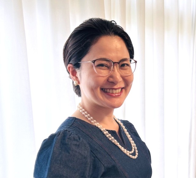 潜在意識コーチ養成講座の体験者の写真(40代女性・元教員)メガネをかけ、紺のワンピースにパールネックレスでクラシックな装いでほほ笑む