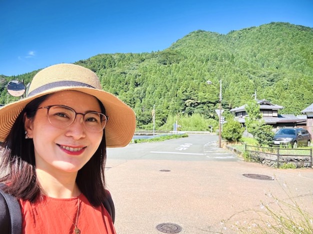 潜在意識コーチ養成講座の体験者の写真(40代女性・元教員)青い空と緑の山を背景に、麦わら帽子をかぶり笑顔いっぱい